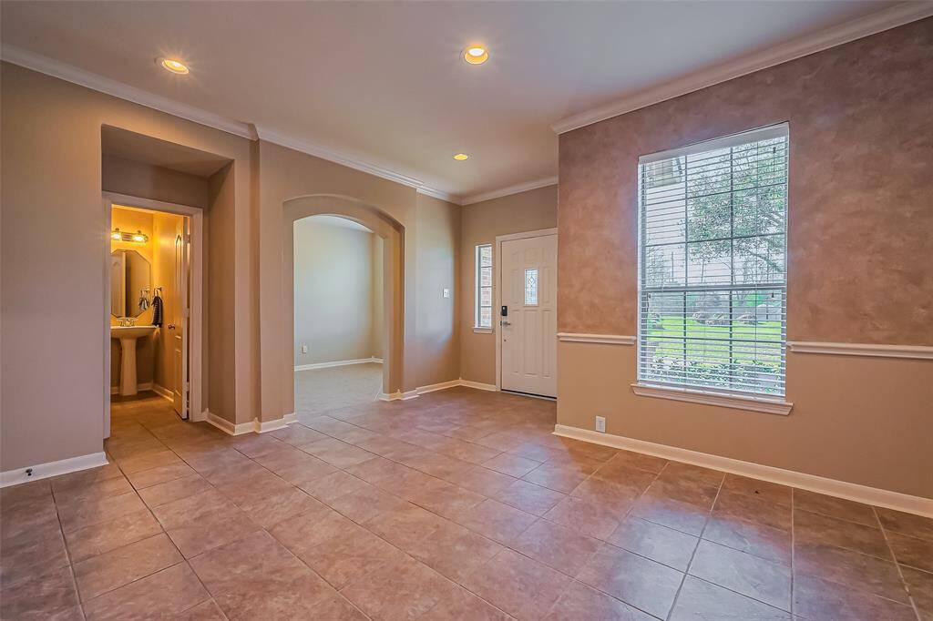 This spacious dining room features neutral tile flooring, soft beige walls, and recessed lighting. It includes a large window with natural light, an arched doorway leading to another area, and a visible entry to a small bathroom with a pedestal sink.