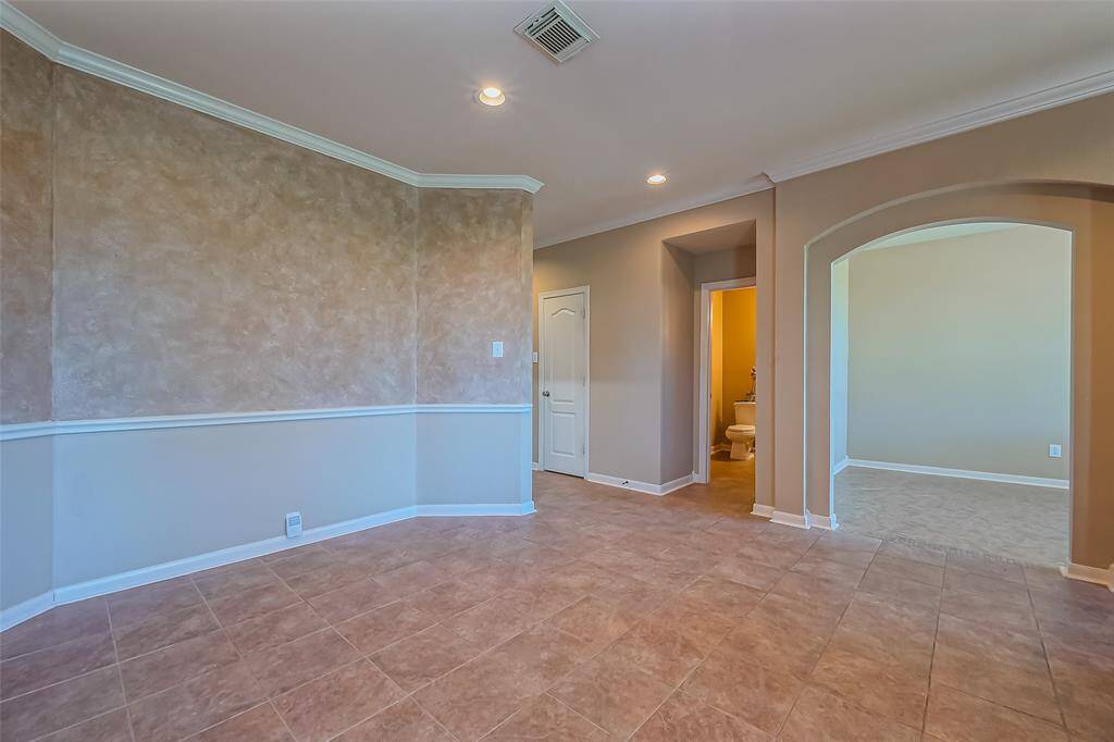 This dining room photo showcases a spacious room with tiled flooring, neutral walls, and crown molding. It features an arched doorway leading to another area and a small hallway with access to a bathroom. The space is bright and inviting, ideal for versatile use.