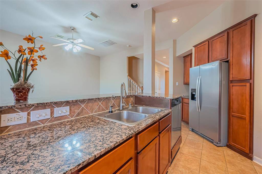 This kitchen features granite countertops, a double sink, and stainless steel appliances, including a large refrigerator. It has warm wood cabinetry, a tile backsplash, and tile flooring.