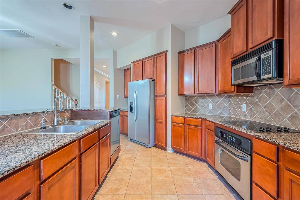 This kitchen features warm wooden cabinetry, granite countertops, and stainless steel appliances, including a double sink and built-in microwave. The tiled backsplash complements the tile flooring, and there's an open view to the adjacent area.