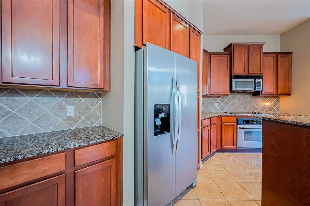 This kitchen features warm wooden cabinetry, granite countertops, and a tiled backsplash. It includes stainless steel appliances like a refrigerator, oven, and microwave, with tile flooring for easy maintenance.
