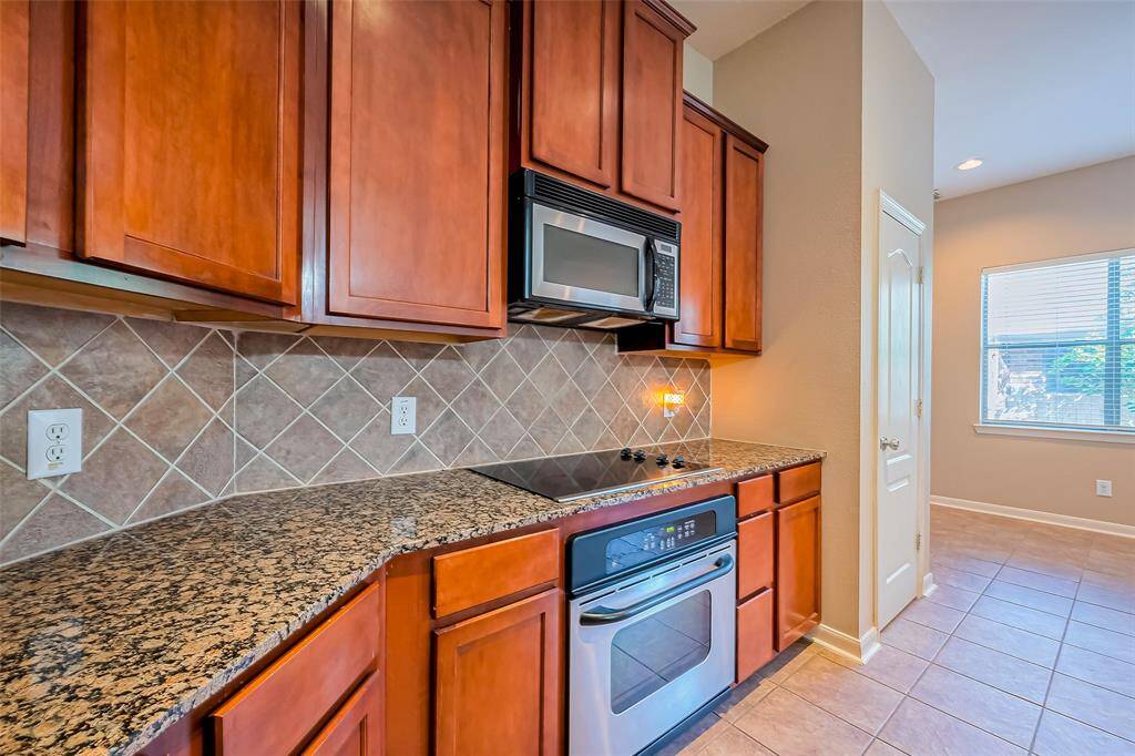 This kitchen features warm wood cabinetry, granite countertops, and a tile backsplash. It includes a built-in microwave and oven, with a ceramic cooktop. The space is well-lit with natural light from a nearby window.