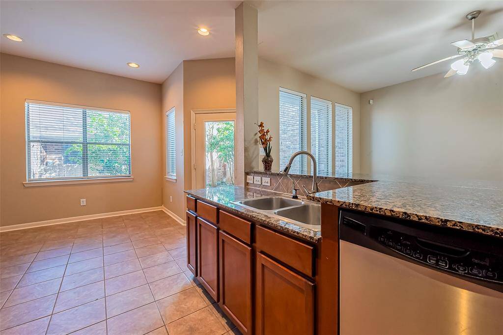 This kitchen features a spacious layout with tile flooring, granite countertops, and a double sink. It includes a dishwasher and ample natural light from large windows, creating a bright and inviting atmosphere.