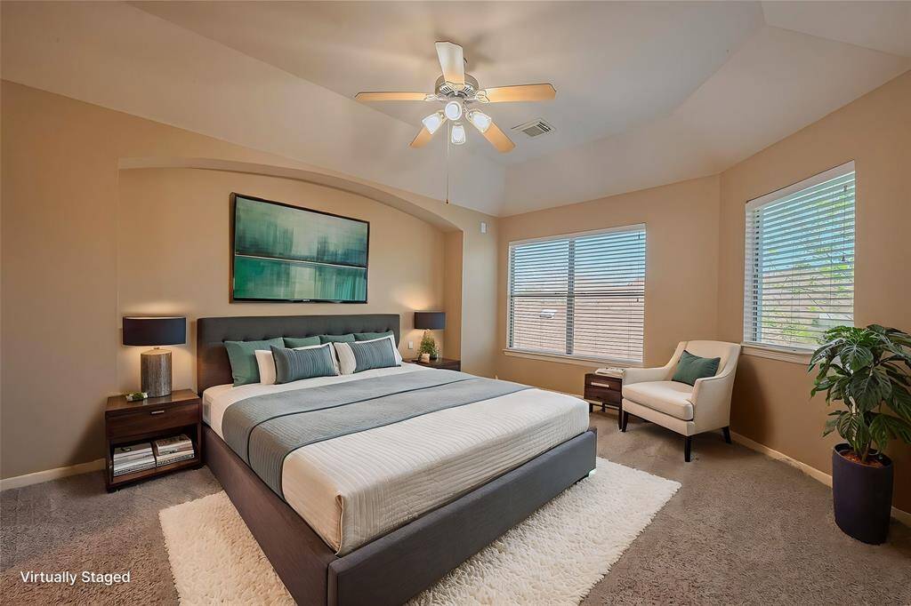 Virtually staged primary bedroom featuring neutral walls and carpeting, abundant natural light from two large windows, and a ceiling fan for added comfort.