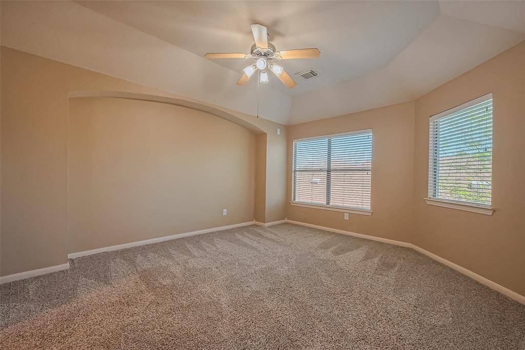 This spacious room features neutral walls, plush carpeting, and ample natural light from multiple windows. It includes a ceiling fan and a unique arched wall design, offering a cozy and inviting atmosphere.