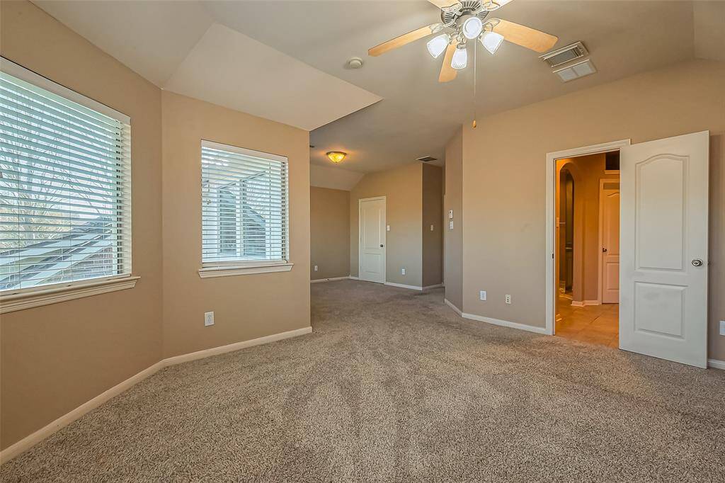 This spacious room features soft, neutral-toned walls, plush carpeting, and an abundance of natural light streaming in through multiple windows. A ceiling fan adds comfort and functionality, while a convenient doorway leads directly to the en suite bath—offering privacy and ease in this cozy, light-filled retreat.