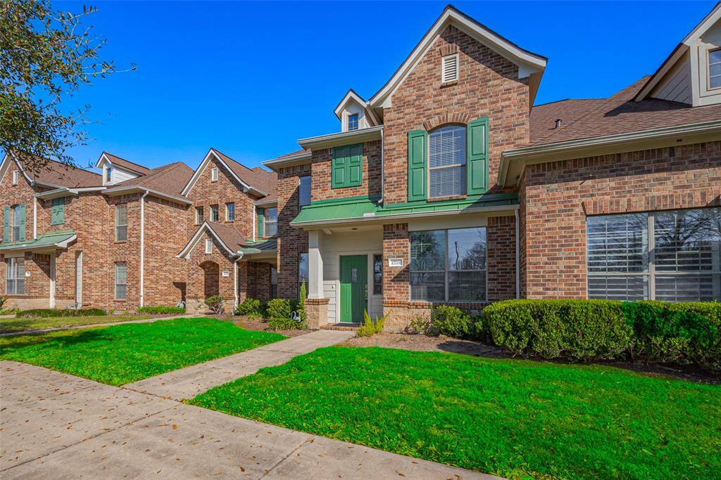 Charming brick townhouse with green shutters and a well-maintained lawn. Features include large front windows, a welcoming entrance, and a neatly paved walkway.