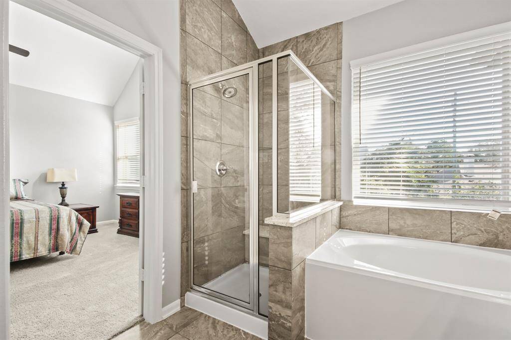 Bright bathroom with stone tile, glass shower, soaking tub, and ample natural light. Connected to a cozy bedroom.