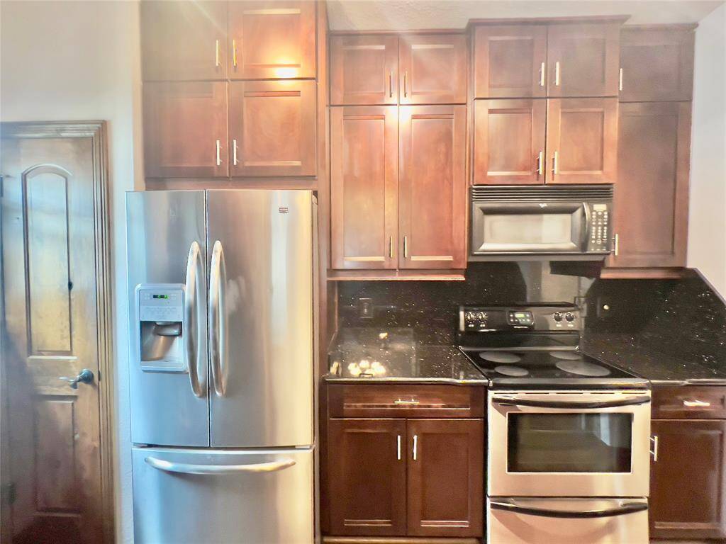 This kitchen features stainless steel appliances, including a double-door refrigerator with a freezer drawer and water dispenser, a microwave above a stove, and dark wood cabinetry. The countertops are black, possibly granite, and there's a pantry door to the left.