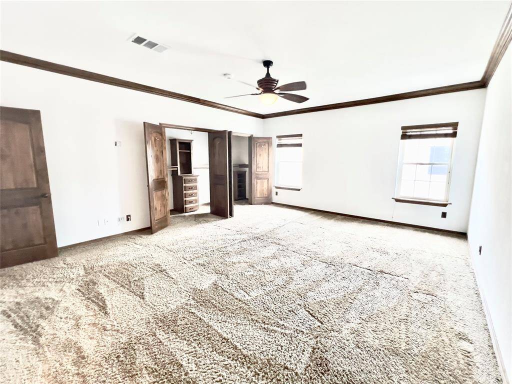 This is a spacious, well-lit room with plush carpeting, featuring a ceiling fan, two windows for natural light, and a built-in shelving unit. The room is complemented by dark wood doors, offering a cozy and elegant feel.