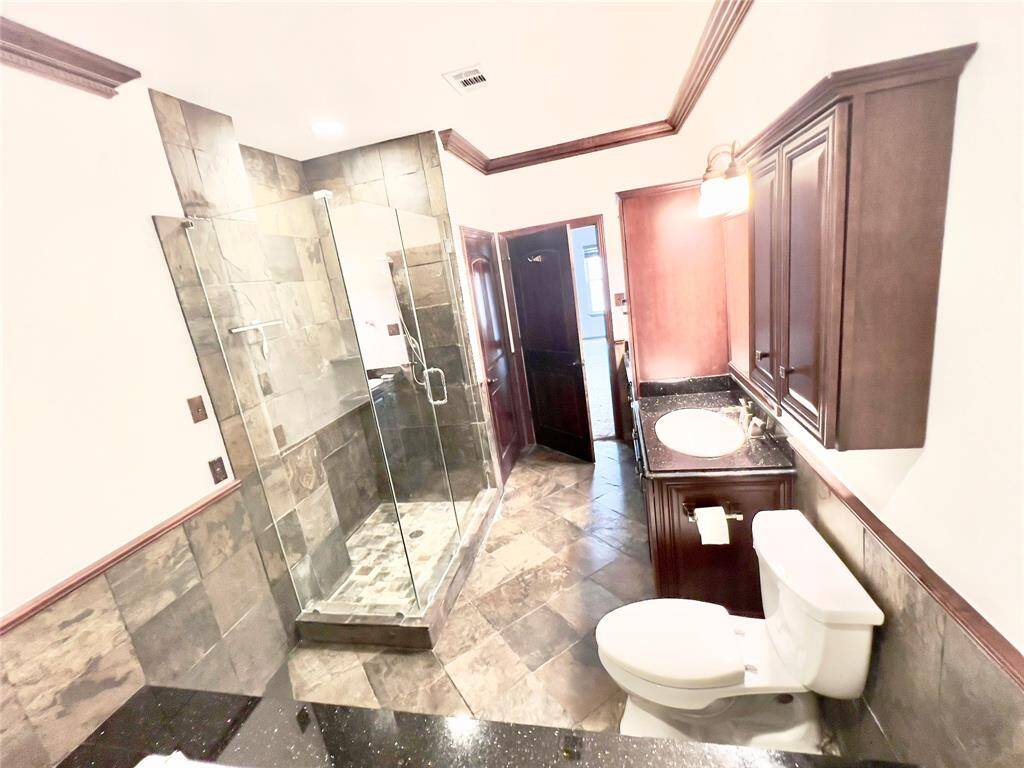 This is a spacious bathroom featuring a large walk-in shower with glass doors, a single vanity with ample cabinetry, and a toilet. The room is finished with sleek, dark stone tiles and has crown molding for an elegant touch.