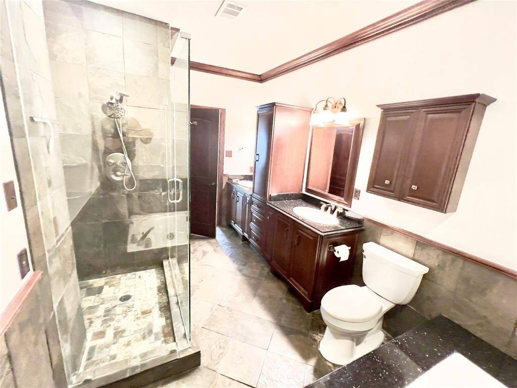 This is a spacious bathroom featuring a large, glass-enclosed shower with stone tiling, a wooden vanity with a dark finish and matching overhead cabinets, and a classic white toilet. The room has crown molding and is illuminated by sconce lighting above the vanity.
