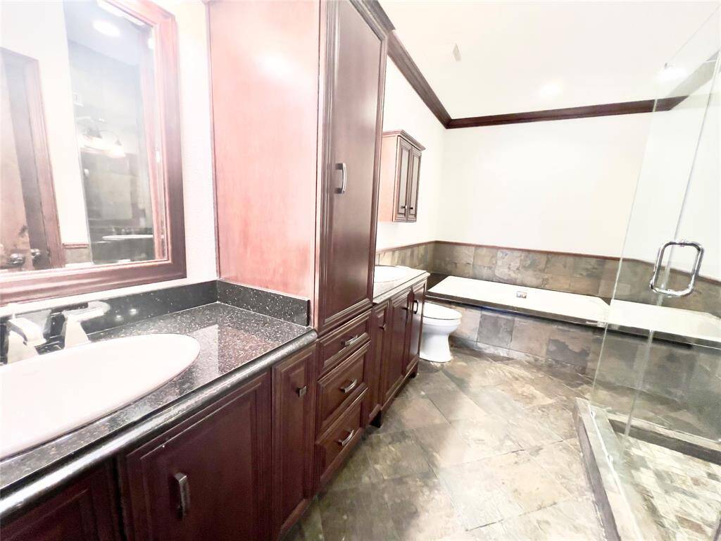 This bathroom features dark wood cabinetry, granite countertops, a large mirror, undermount sink, toilet, and a spacious walk-in shower with glass enclosure next to a built-in bathtub. The room has tile flooring and partial tile walls, creating a cohesive and elegant look.