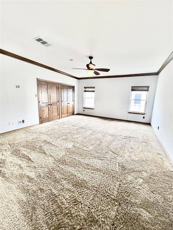 This is a spacious room with plush carpeting, natural light from two windows, a ceiling fan, and a set of wooden double doors. The walls are painted white with dark trim, giving it a clean and classic look.