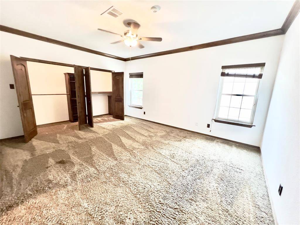 This is a spacious room with plush carpeting, featuring a ceiling fan, two windows for natural light, and built-in shelving units. It has a neutral color palette and wood trim.