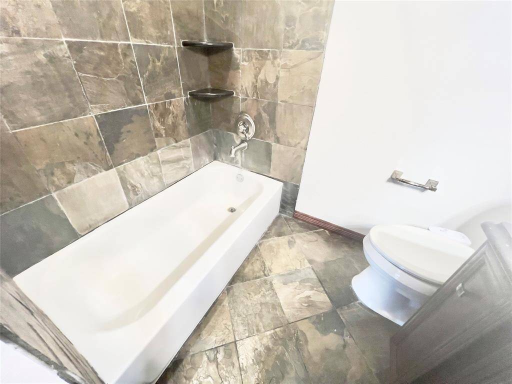 This is a bathroom featuring a built-in bathtub with earth-toned tile surround, a toilet, and similar tiled flooring. There are also built-in shelves for storage within the tiling. The space appears modern and well-maintained.