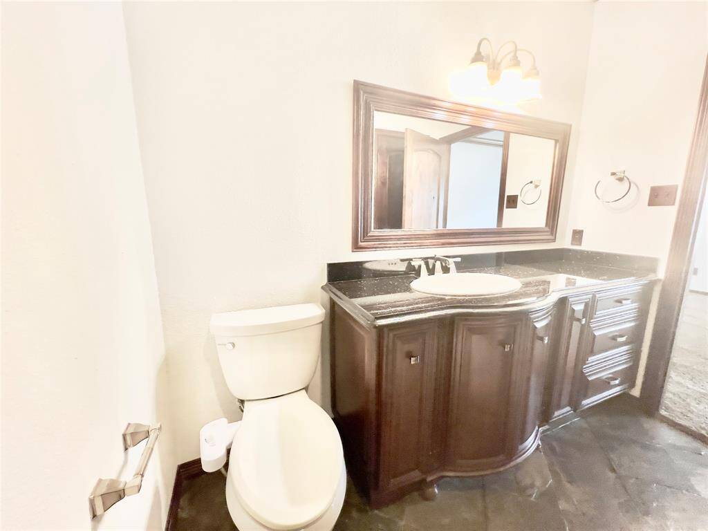 This is a bathroom featuring a wood-finished vanity with a granite countertop and an undermount sink, a large framed mirror, and a toilet. The space has tiled flooring and is well-lit.