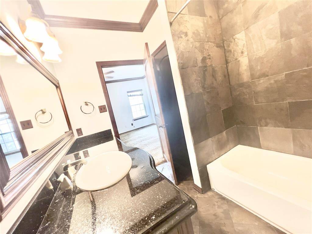 This is a modern bathroom with a dark granite countertop, an undermount sink, large mirrors, stylish wall sconces, and a full bathtub with dark tile surround. It has a warm, neutral color palette and a doorway leading to an adjacent room.