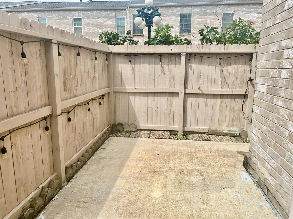 This photo shows a small, private outdoor patio area with a high wooden fence for privacy and string lights for ambiance. The space is paved and bordered by a residential building on one side, suggesting a cozy spot for personal use.