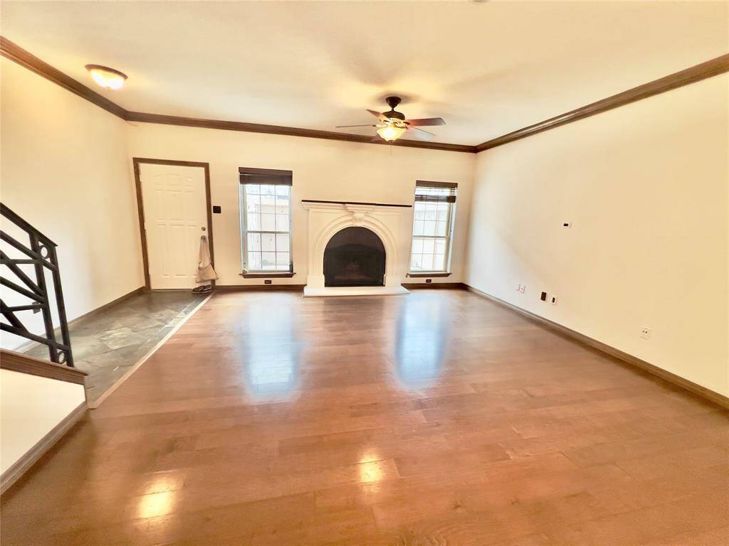 This is a spacious, well-lit room with hardwood floors, featuring a classic fireplace, large windows, and an entry door. There’s also a staircase leading to another level, suggesting a multi-story home.