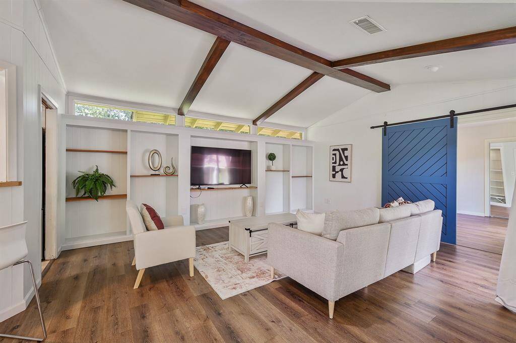 Spacious and full of character, this living area features wooden beams that beautifully contrast with the crisp white ceiling and walls. A charming blue barn door adds personality, while a built-in shelf spans one wall, serving as both functional storage and a striking focal point. Open to the dining area yet maintaining a distinct sense of space, the room also connects effortlessly to the kitchen through an open doorway.