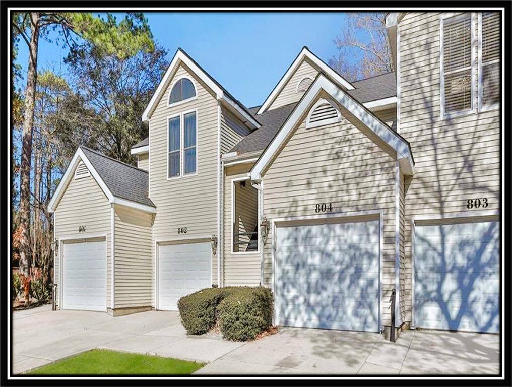 This photo shows a row of modern condos with beige siding and white garage doors. Each unit has a pitched roof and large windows. The area is surrounded by trees, providing a serene and inviting atmosphere.