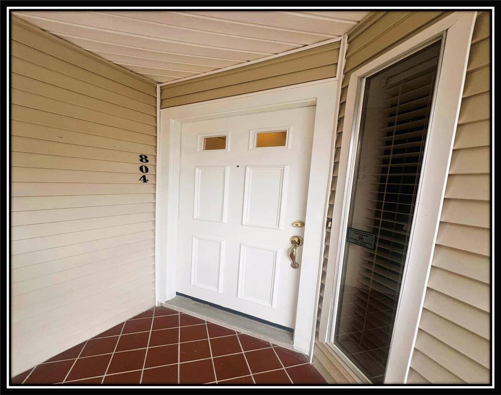 This photo shows a welcoming front entrance with a white door featuring small windows, a brass handle, and a sidelight window. The exterior has light beige siding and a brick-patterned tile floor, creating a neat and inviting entryway.