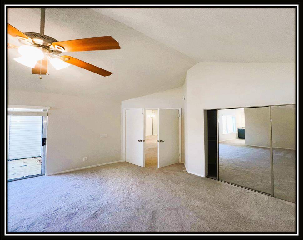 Bright, spacious bedroom with vaulted ceilings, a ceiling fan, mirrored closet doors, and a sliding glass door leading to an outdoor area. Neutral carpeting and walls create a clean, inviting atmosphere.