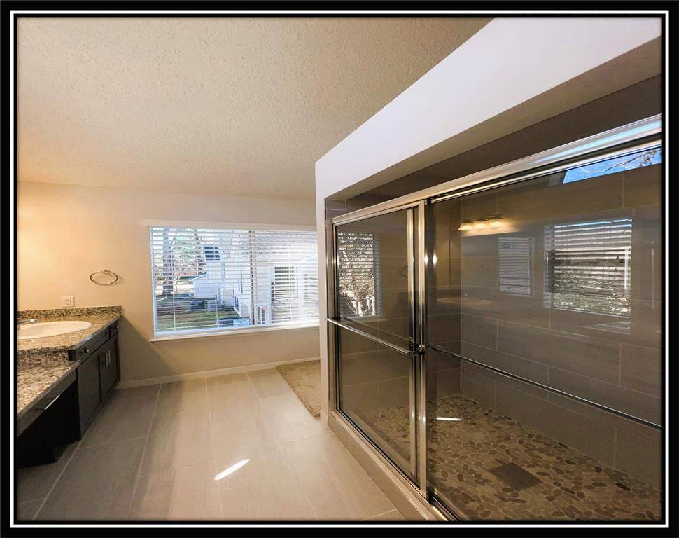 This modern bathroom features a spacious, glass-enclosed shower with pebble flooring, a granite countertop with double sinks, and a large window with blinds that allows natural light to brighten the space.