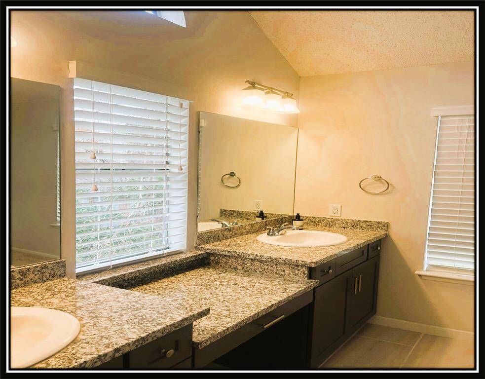 This photo showcases a modern bathroom with a spacious granite countertop, dual sinks, large mirrors, and ample natural light from the windows with blinds.