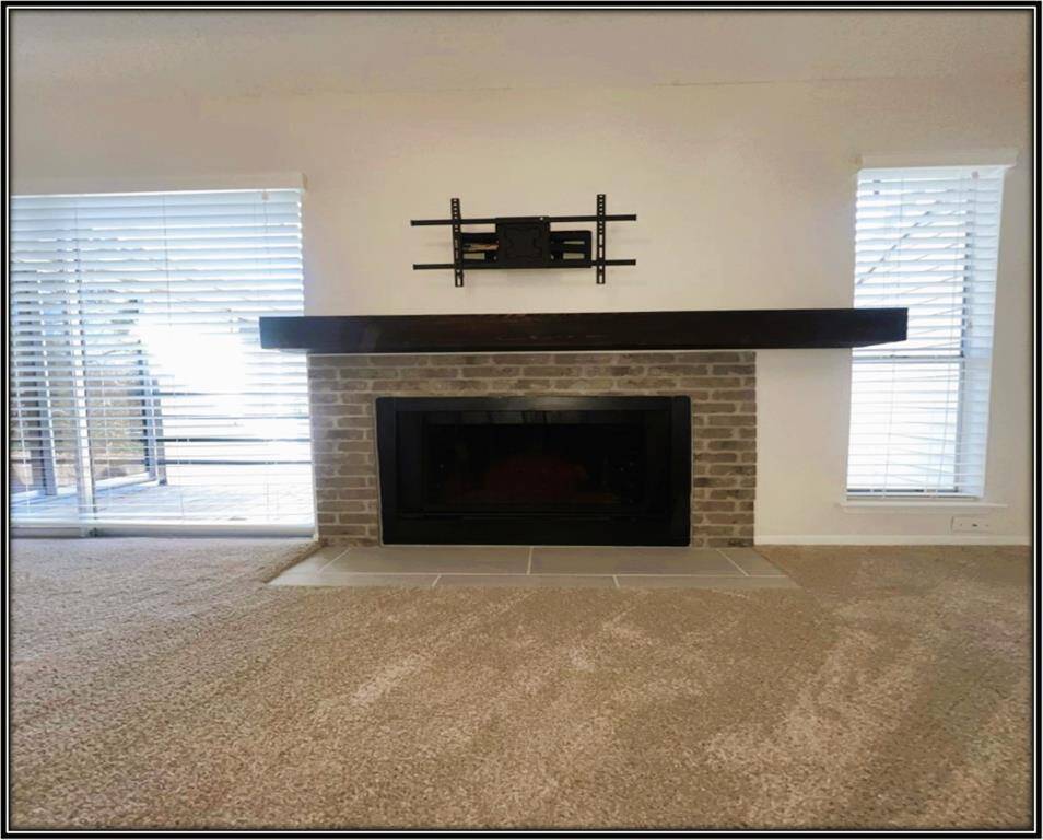 This living room features a cozy brick fireplace with a dark wood mantel, flanked by large windows with blinds, allowing plenty of natural light. The room is carpeted, creating a warm and inviting atmosphere.