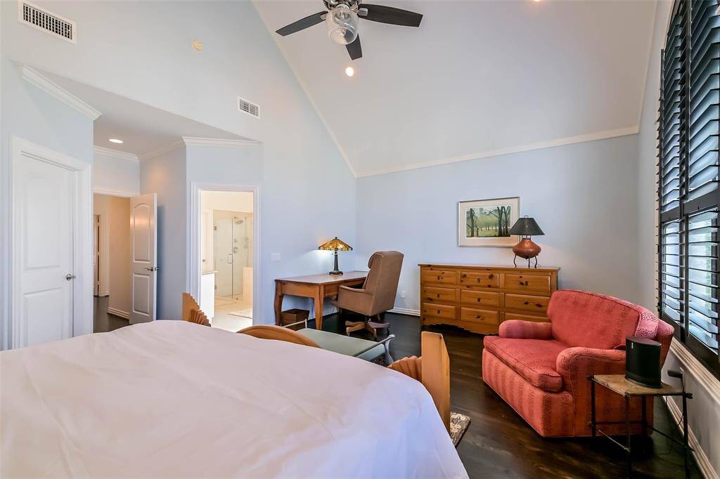 This spacious bedroom features high ceilings and ample natural light, with a large window and modern ceiling fan. It includes a cozy sitting area, wooden furniture, and an ensuite bathroom for convenience.