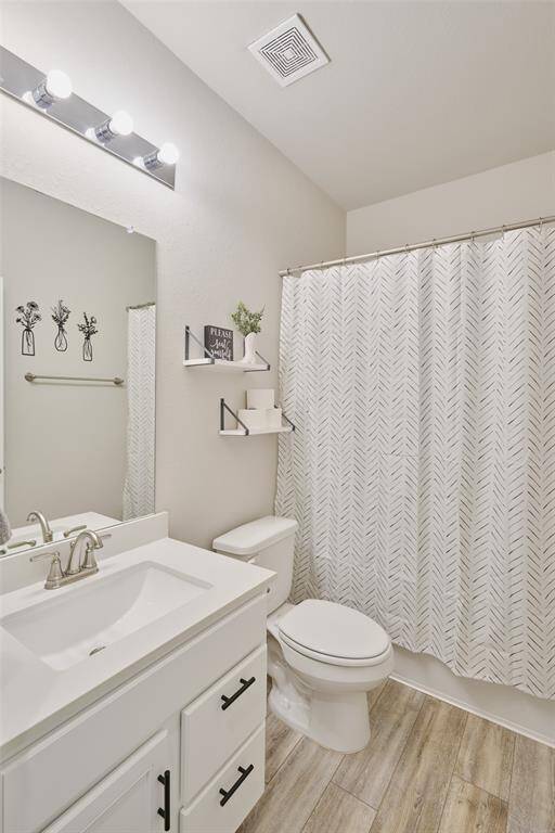 This bathroom features a modern design with light wood flooring, a sleek white vanity, and a large mirror. It includes a shower with a patterned curtain, tasteful wall decor, and floating shelves for added storage and style.