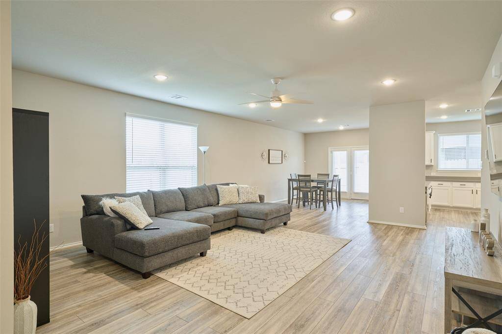 This photo showcases a spacious, open-concept living area with light wood flooring and neutral walls. It features a cozy gray sectional sofa, a dining area with a table and chairs, and a modern kitchen with white cabinets. The room is well-lit with natural light from large windows and recessed ceiling lights.