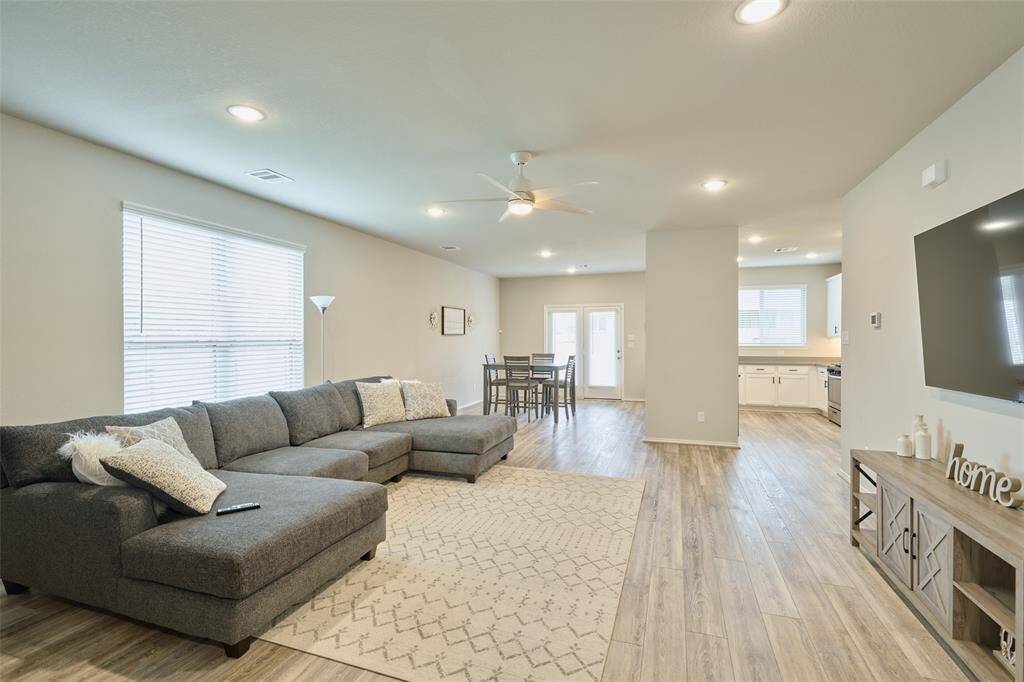 This spacious living area features an open floor plan with a cozy sectional sofa, modern flooring, and ample natural light. The room flows into a dining area and a bright kitchen, creating a welcoming and functional space for family living.