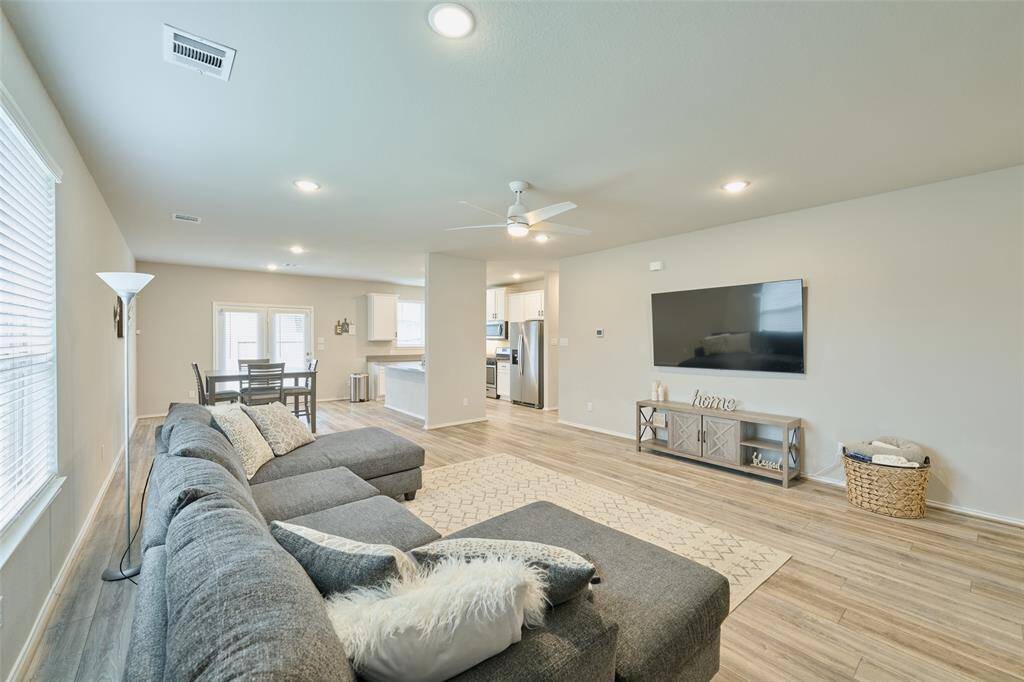 This photo showcases a spacious, open-concept living area with modern decor. It features a large sectional sofa, wall-mounted TV, and a dining area adjacent to the kitchen. The space is well-lit with natural light from multiple windows, and has light wood flooring and neutral walls.