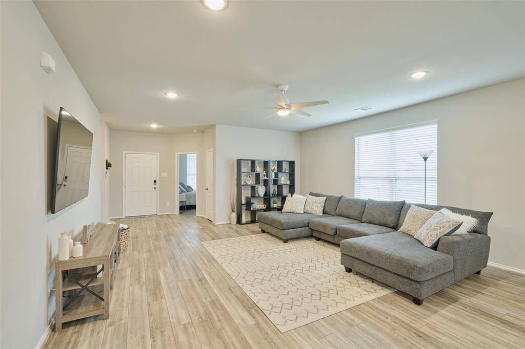 This spacious, well-lit living room features a large sectional sofa, modern decor, and wood-style flooring. It includes a wall-mounted TV, ceiling fan, and ample natural light from a large window. The open layout offers easy access to adjacent rooms.