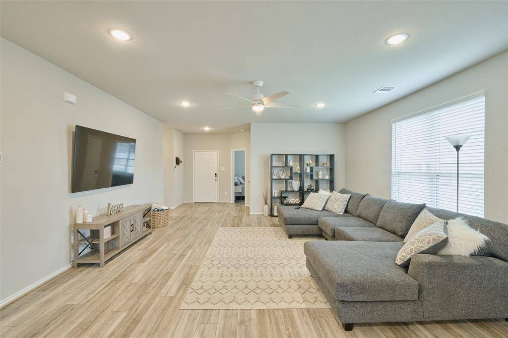 Spacious living room with light wood flooring, neutral walls, and ample natural light. Features a large sectional sofa, wall-mounted TV, and modern shelving. Perfect for relaxation and entertainment.