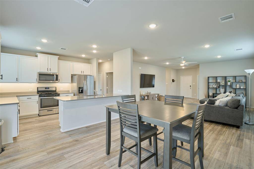 This photo shows an open-concept living space featuring a modern kitchen with stainless steel appliances and white cabinetry. The dining area has a sleek table, and the cozy living room offers a comfortable seating setup with a wall-mounted TV. The space is bright and airy with wood-style flooring throughout.