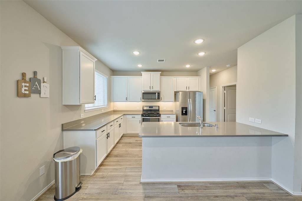This kitchen features modern white cabinetry, stainless steel appliances, and a spacious island with a sink. The open layout and ample lighting create a bright, inviting space with light wood flooring.