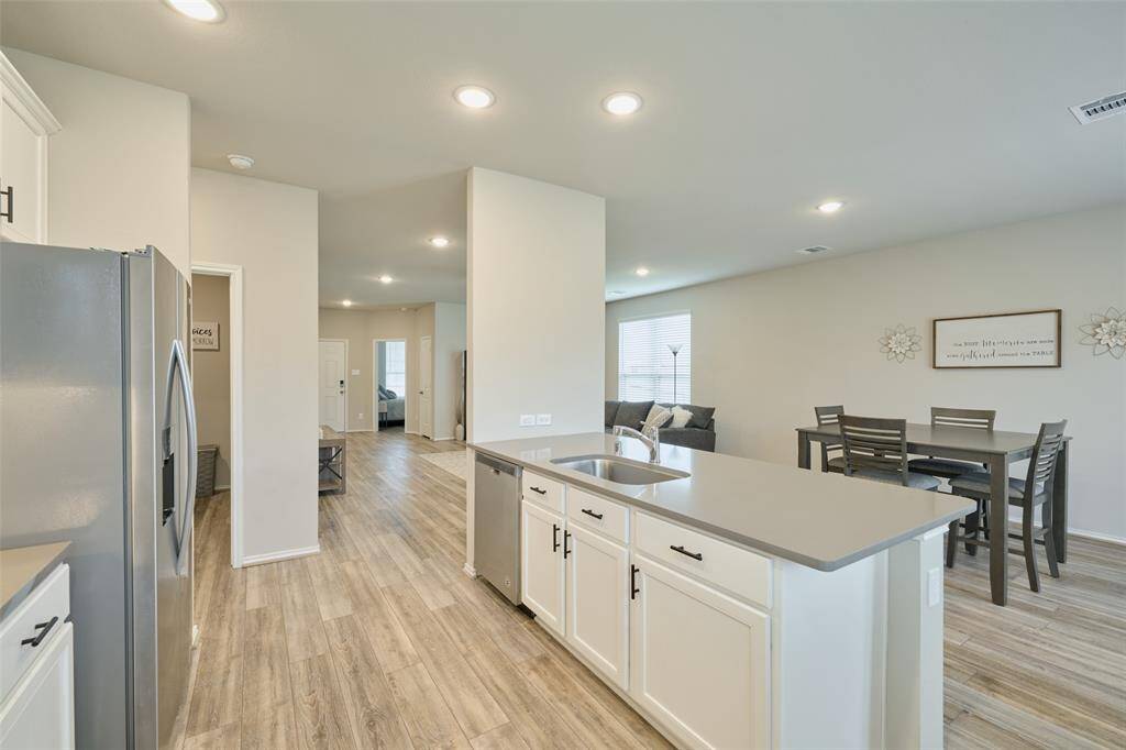 This photo shows a bright, open-concept kitchen and living area featuring light wood flooring, modern white cabinetry, stainless steel appliances, and a spacious island. The adjacent dining area and living space offer ample room for relaxation and entertaining.