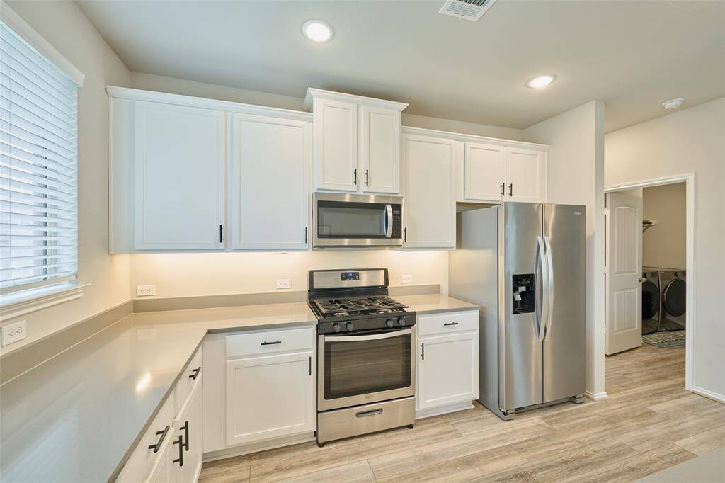 Modern kitchen with stainless steel appliances, light wood flooring, and ample white cabinetry. Features a gas stove, microwave, and large fridge. Bright and spacious with natural light from a window. Adjacent to a laundry area.