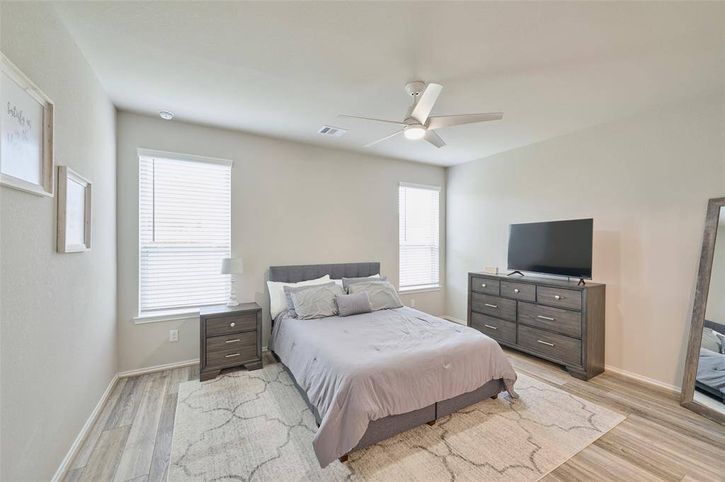 This bedroom features a modern design with light-colored walls and wood flooring. It includes a cozy bed with a gray headboard, matching nightstands, and a dresser with a TV. The room is well-lit with natural light from two windows and has a ceiling fan for comfort.