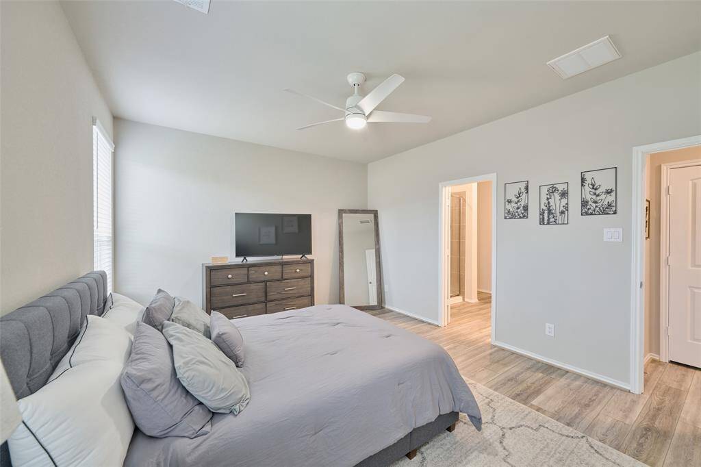 This bedroom is bright and spacious, featuring a large bed, wood-style flooring, and neutral walls. It includes a ceiling fan, a dresser with a TV, and an adjacent bathroom for convenience.