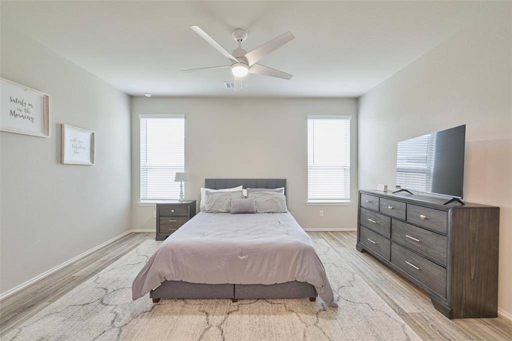 This bedroom features a modern design with neutral tones, a comfortable bed, and ample natural light from two large windows. It includes a ceiling fan, a stylish dresser with a TV, and elegant decor for a serene atmosphere.