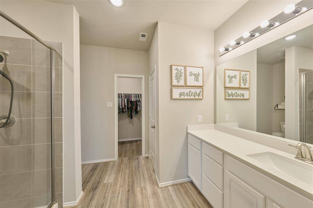 This modern bathroom features a spacious walk-in shower with sleek tiling, a large vanity with ample counter space and lighting, and an adjoining walk-in closet. The neutral tones and wood-style flooring create a warm and inviting atmosphere.