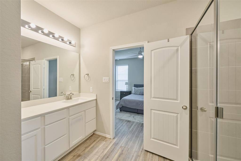 This photo showcases a bright, modern bathroom with a large vanity, ample storage, and a sleek glass shower. The room features neutral tones and opens into a cozy bedroom with wooden flooring.