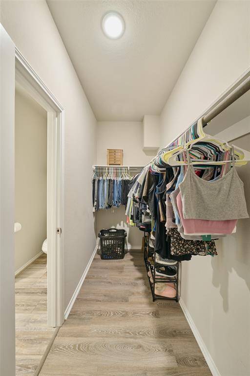 This photo shows a spacious walk-in closet with wooden flooring, equipped with hanging rods and shelves for clothing. It features a ceiling light and has an adjacent door leading to another room.