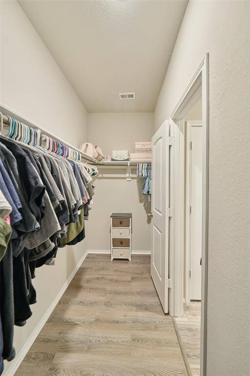 This is a spacious walk-in closet with light wood flooring and neutral walls. It features ample hanging space on both sides, a shelf for storage, and a small cabinet at the end. Ideal for organizing clothing and accessories.