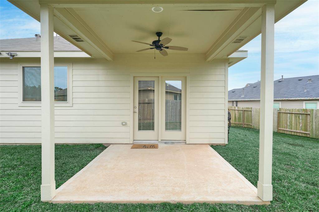 This photo shows a cozy backyard patio with a covered area, ceiling fan, and concrete flooring. It features a welcoming door with blinds, a small doormat, and is surrounded by a fenced grassy yard, perfect for outdoor relaxation.